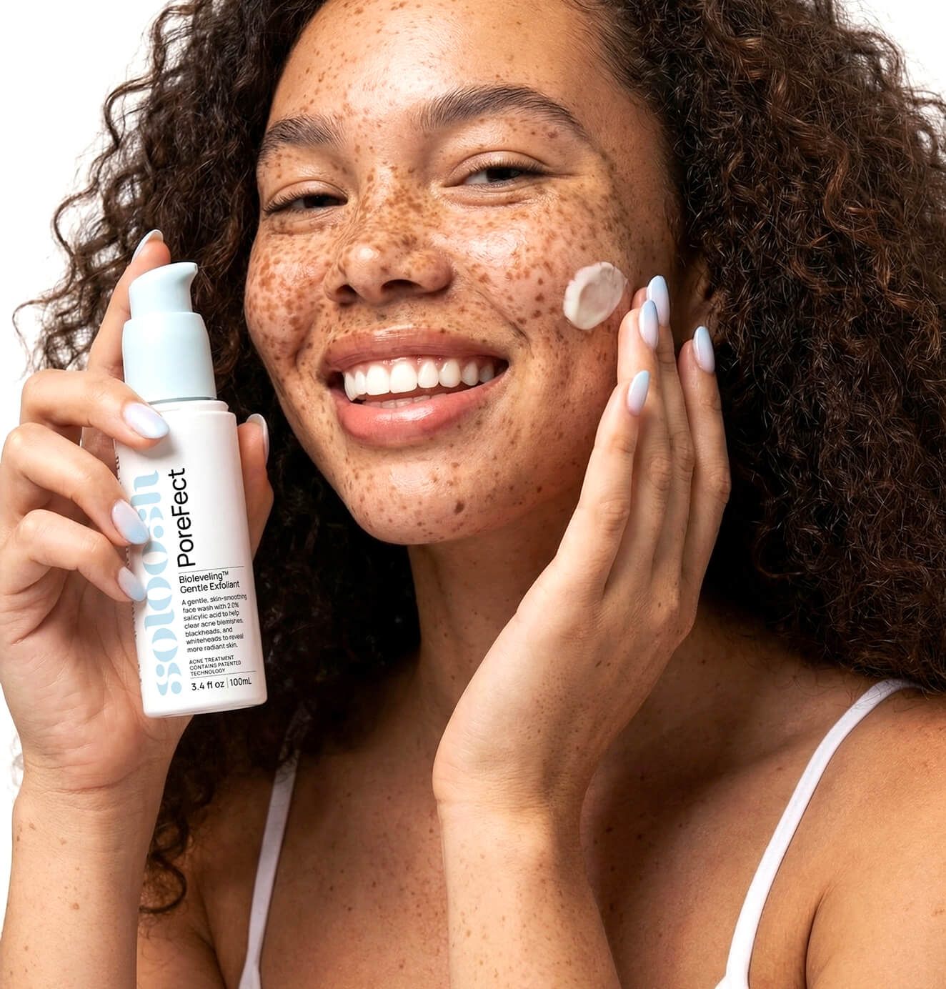 Smiling woman with freckles applying skincare cream while holding Soloosh Porefect exfoliant bottle.