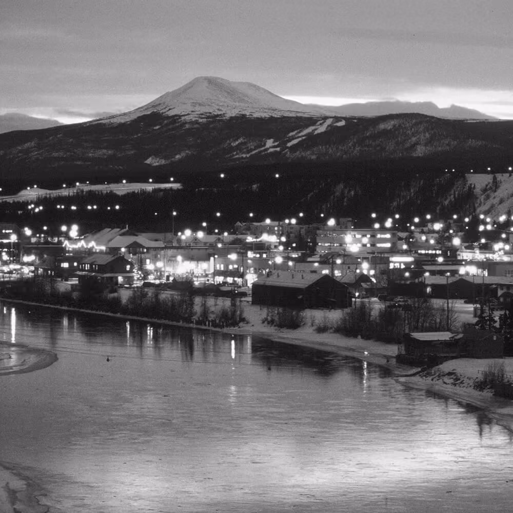 Whitehorse waterfront at dusk with snowy mountain backdrop and glowing city lights.