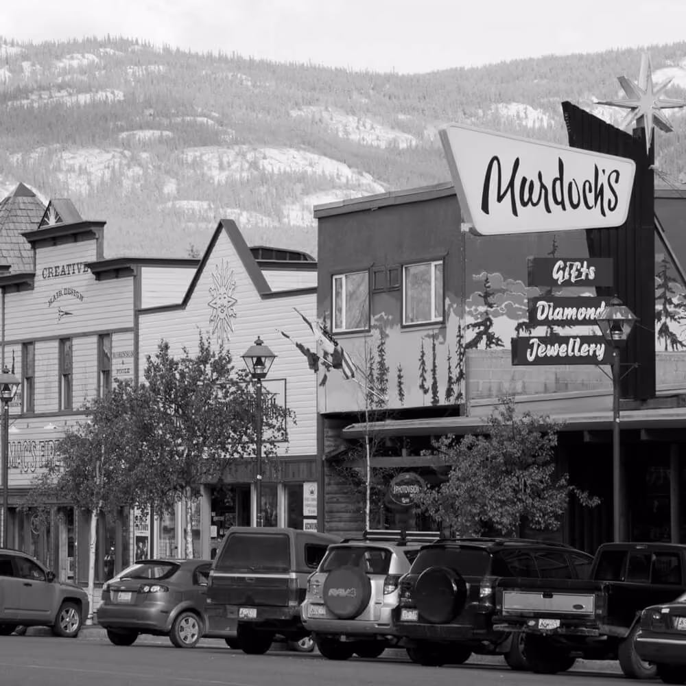 Downtown Whitehorse streetscape with local shops, parked vehicles, and mountain scenery.