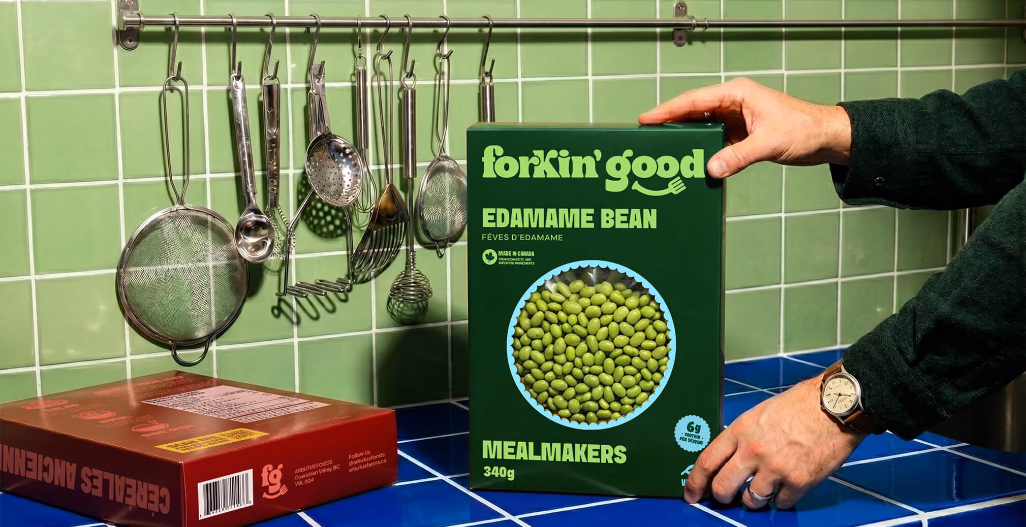 Hand placing a Forkin’ Good Edamame Bean Mealmakers box onto a blue tiled countertop beside hanging kitchen utensils against a green tiled wall, showcasing bold food packaging and playful branding.