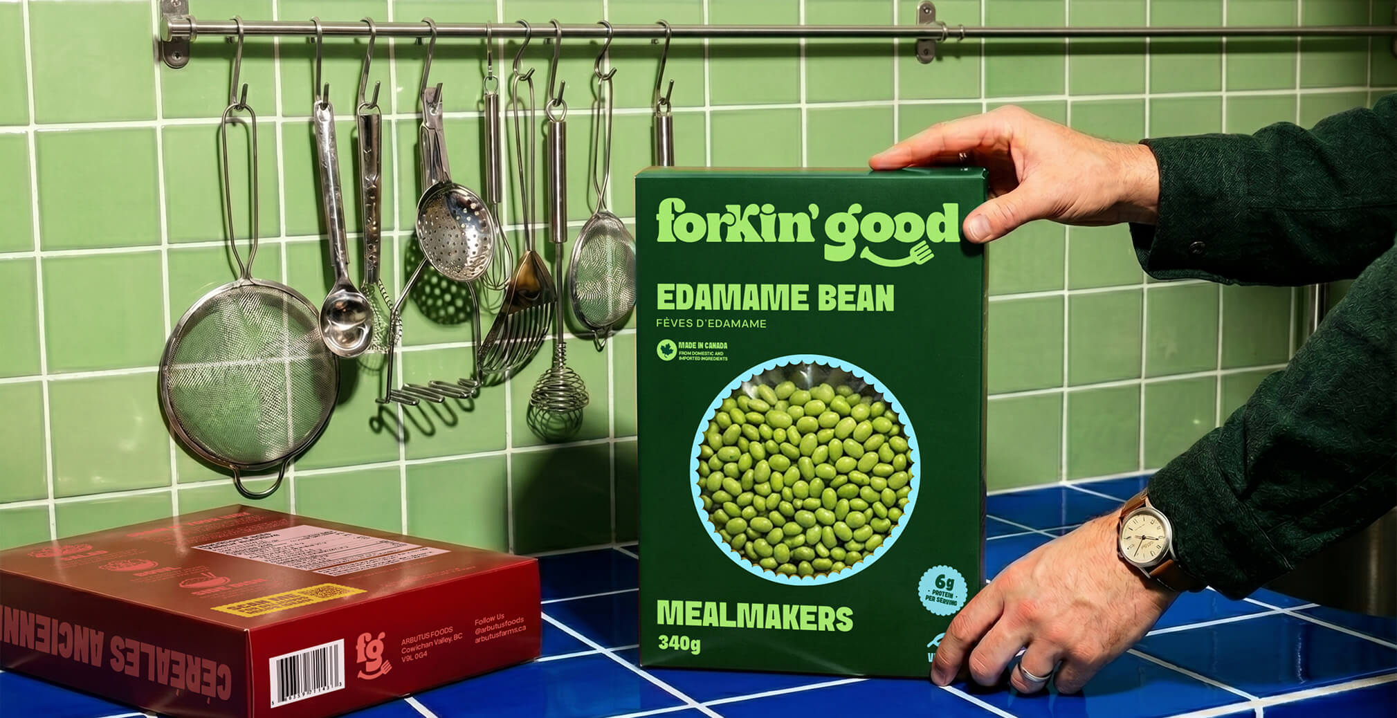 Hand placing a Forkin’ Good Edamame Bean Mealmakers box onto a blue tiled countertop beside hanging kitchen utensils against a green tiled wall, showcasing bold food packaging and playful branding.
