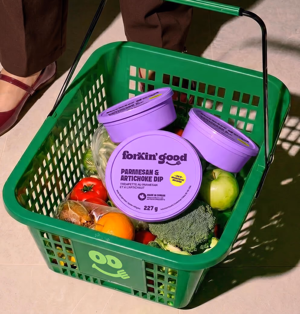 Green grocery basket with fresh produce and Forkin’ Good Parmesan & Artichoke Dip, showcasing retail packaging and food branding.
