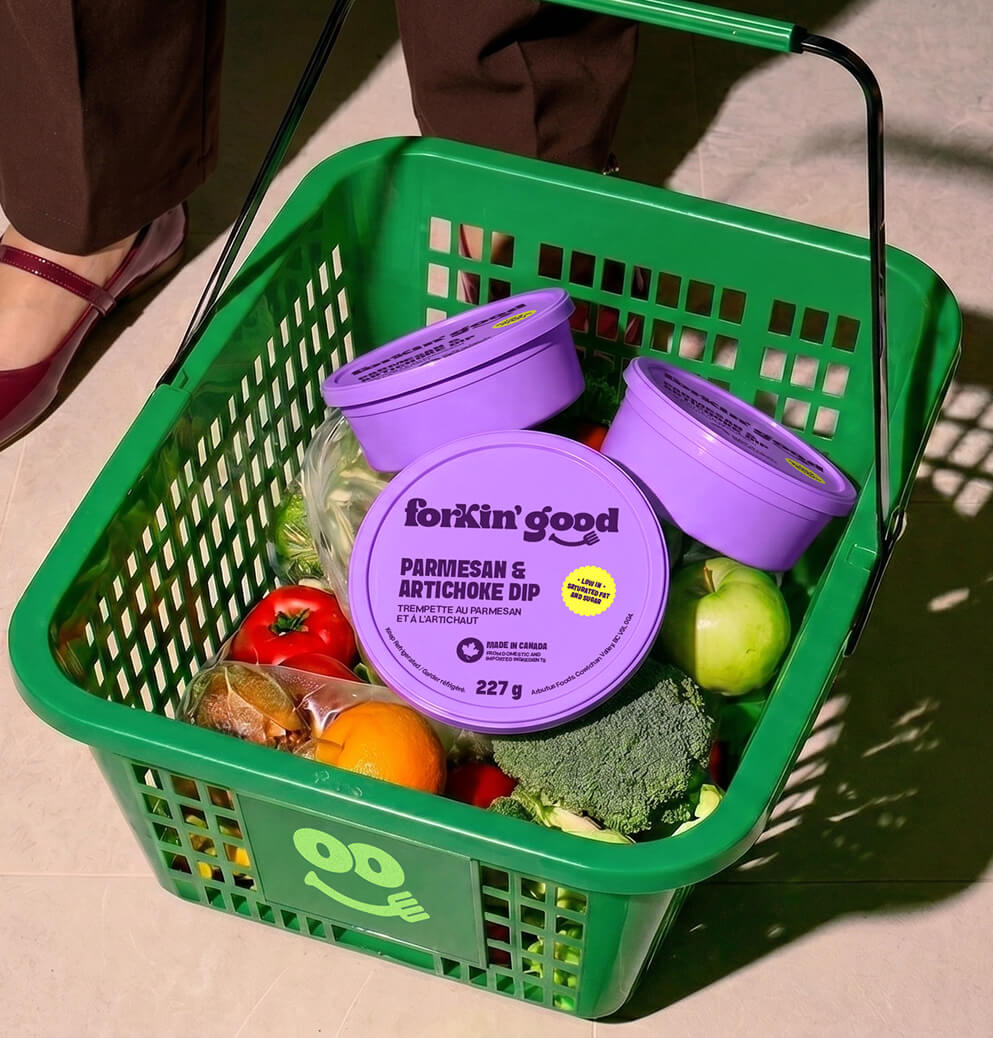 Grocery basket filled with vegetables and Forkin’ Good Parmesan & Artichoke Dip containers, showcasing vibrant retail packaging and lifestyle branding.
