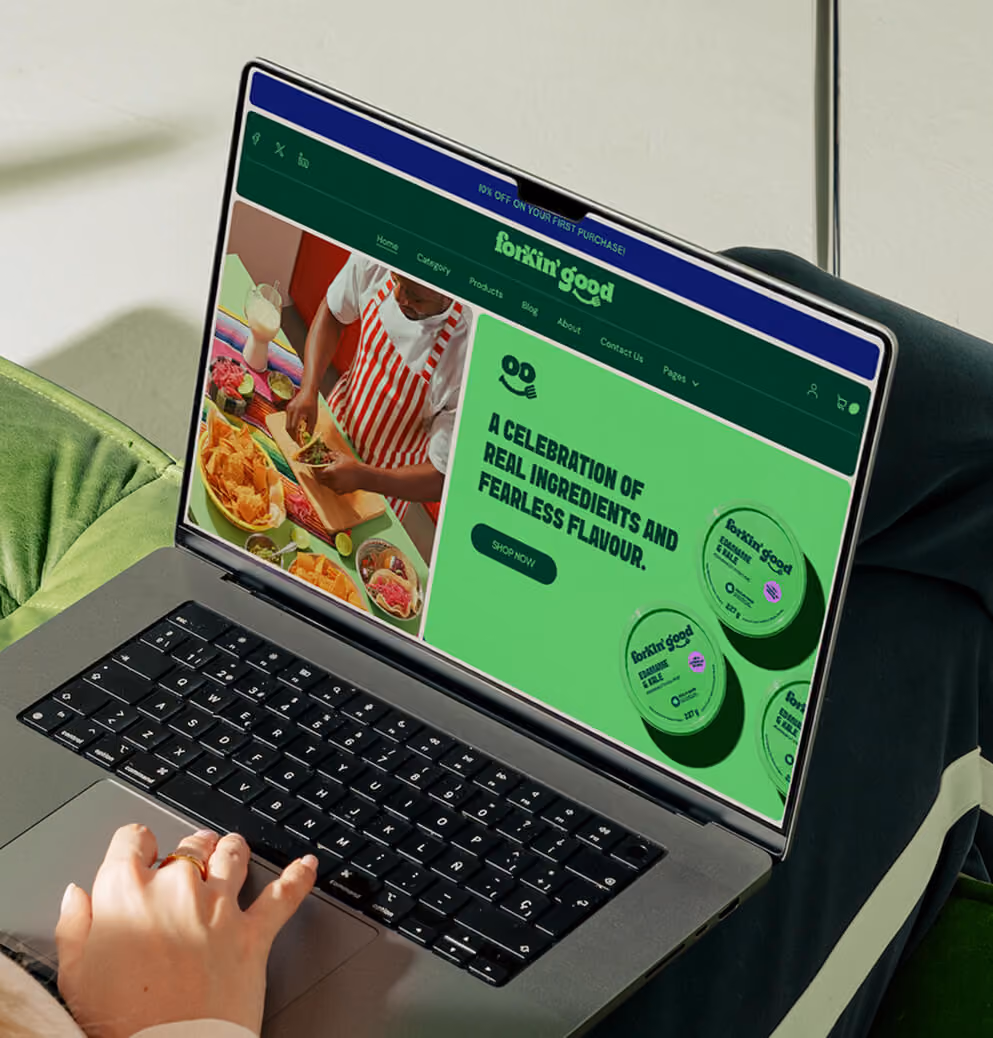 Laptop displaying the Forkin’ Good website homepage with vibrant green branding and food product imagery in a cozy modern interior setting.