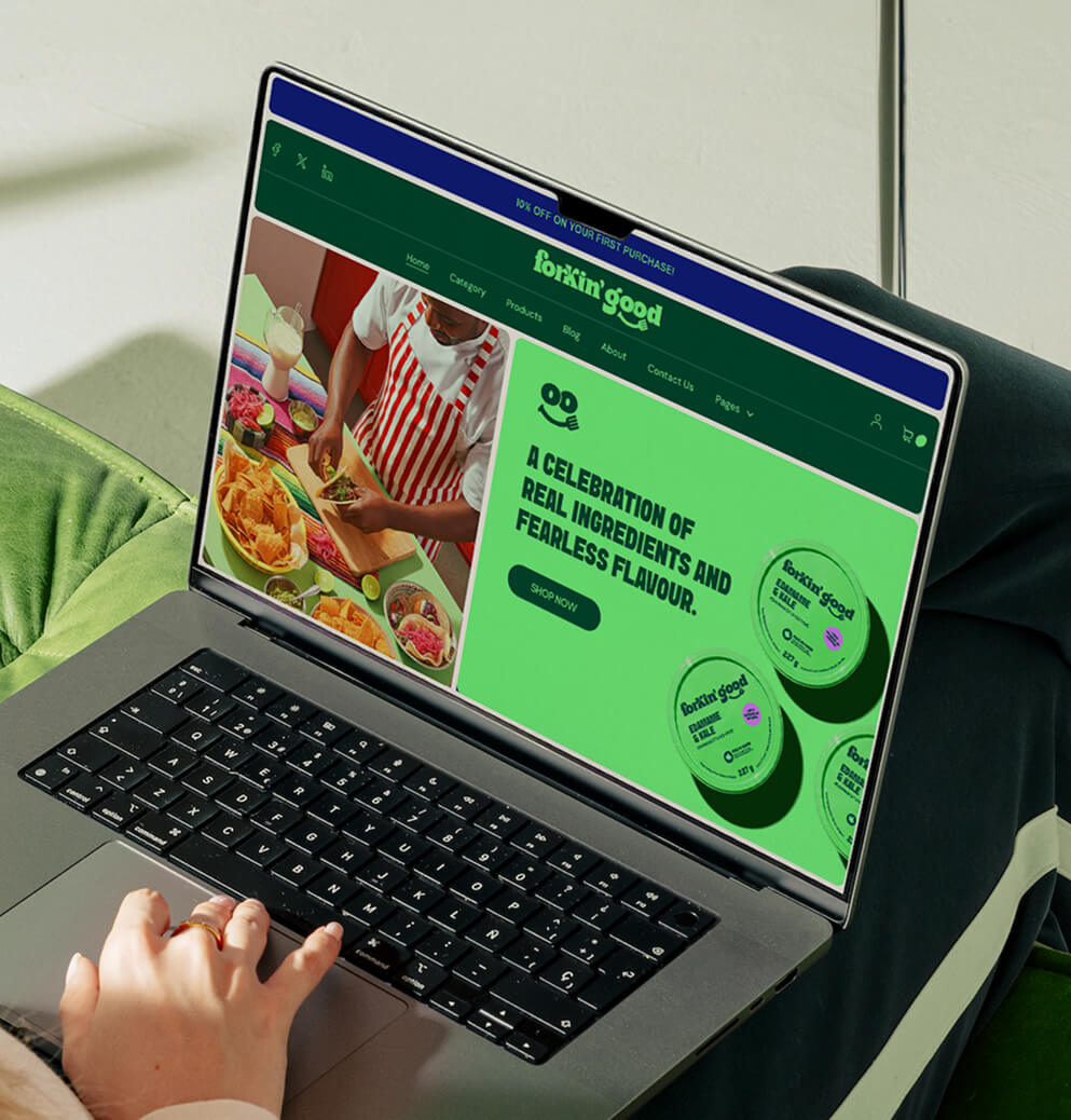 Laptop displaying the Forkin’ Good website homepage with vibrant green branding and food product imagery in a cozy modern interior setting.