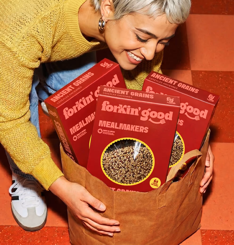 Smiling person holding a grocery bag filled with Forkin’ Good Ancient Grains Mealmakers boxes in a warm-toned lifestyle food branding photoshoot.