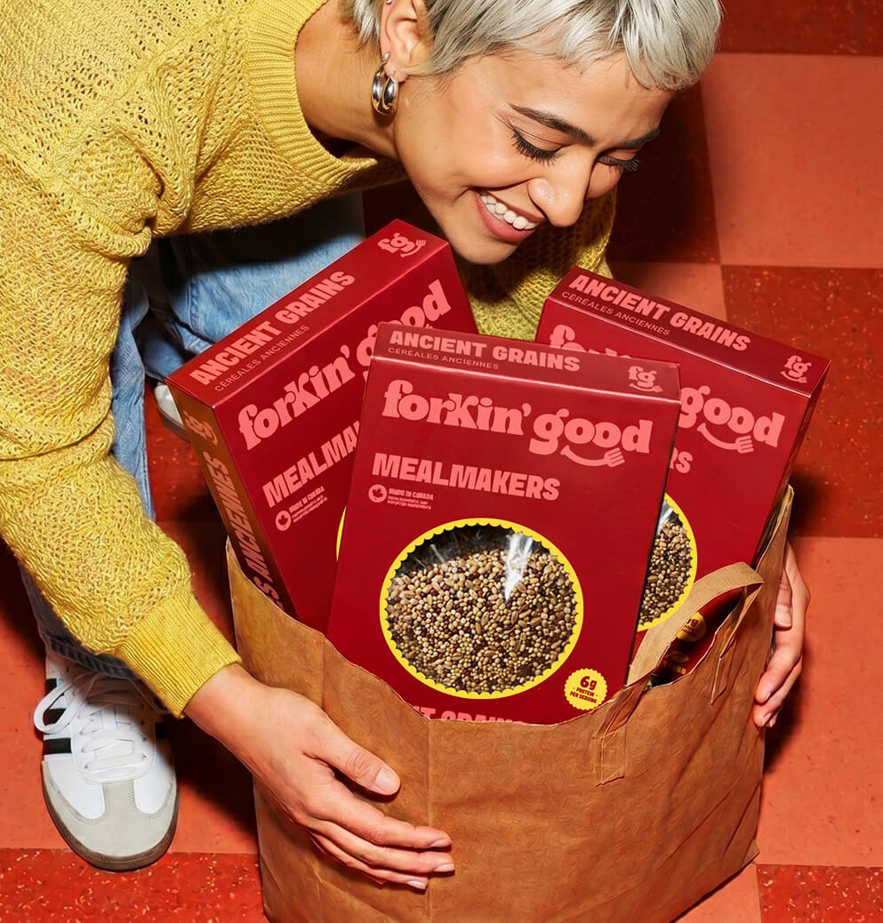 Smiling person holding a grocery bag filled with Forkin’ Good Ancient Grains Mealmakers boxes in a warm-toned lifestyle food branding photoshoot.