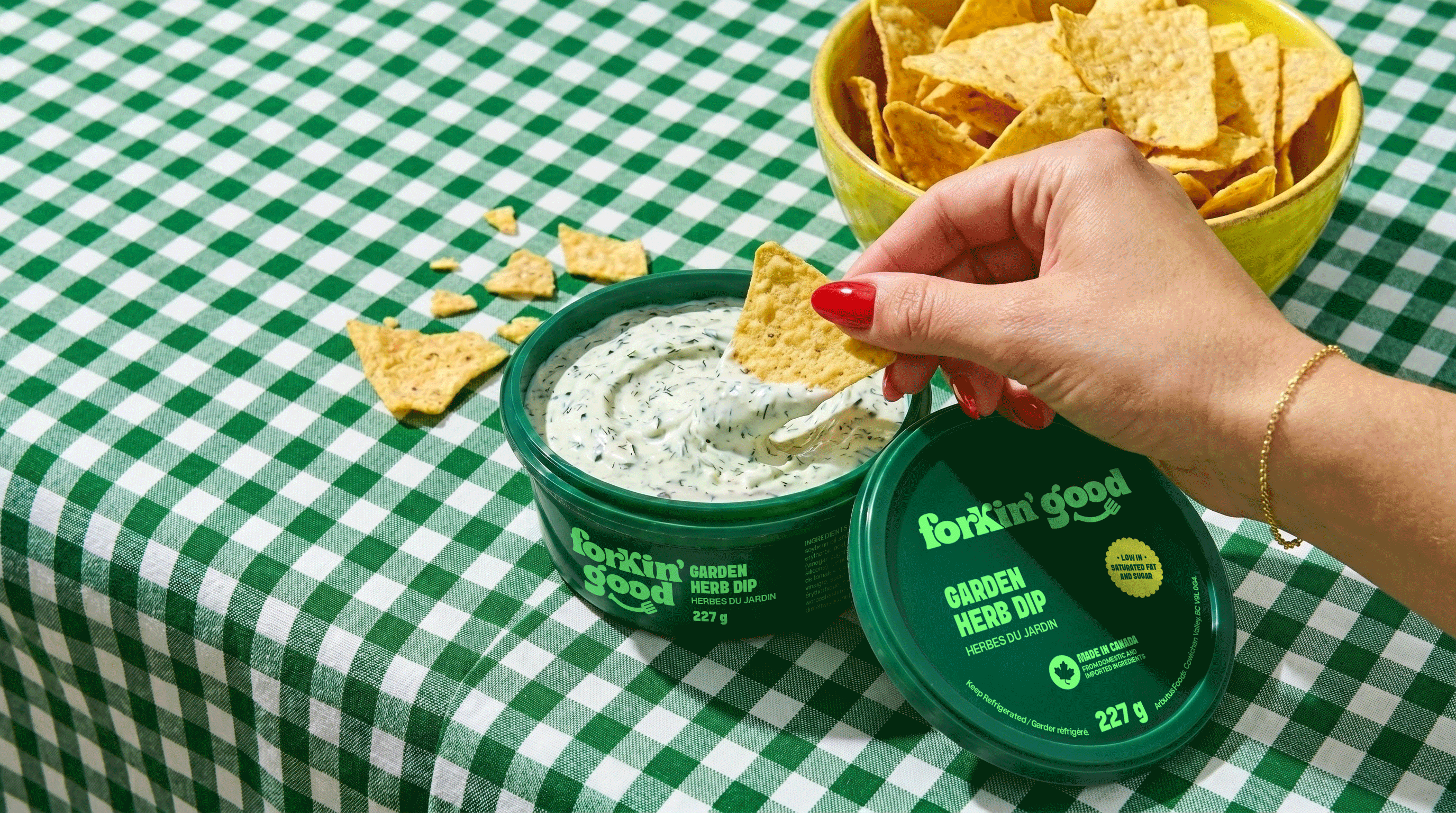 Lifestyle product photo featuring a hand dipping a tortilla chip into Forkin’ Good Garden Herb Dip on a green checkered tablecloth, with branded packaging and a bowl of chips creating a fresh, casual snack scene.