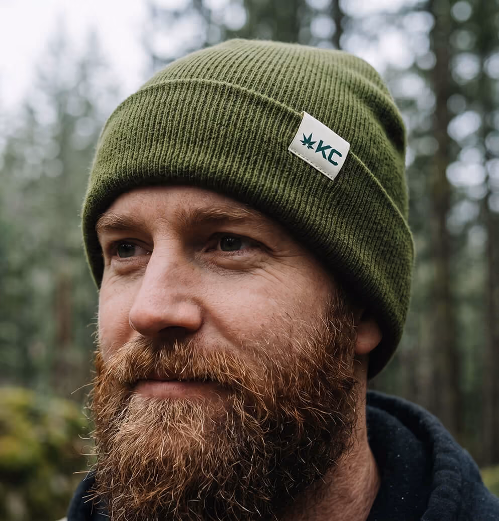 Portrait of a bearded man wearing a green Koots Canna beanie outdoors in a forest setting.