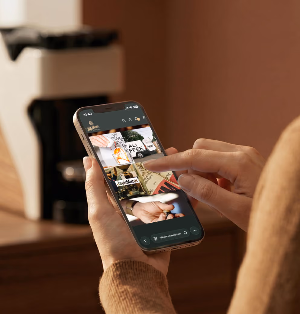 Person browsing the Milton Coffee website on a smartphone with the coffee machine featured in the background, highlighting digital brand experience and e-commerce design.
