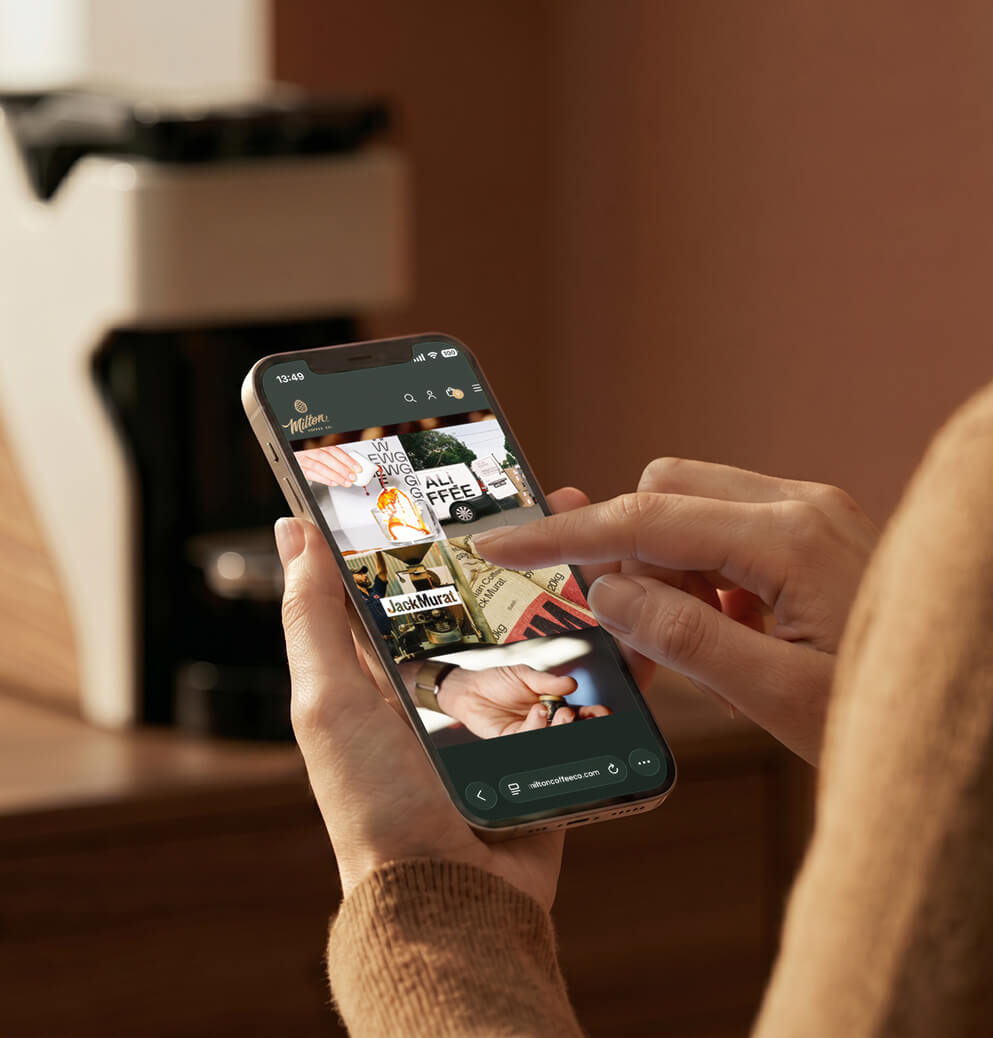 Person browsing the Milton Coffee website on a smartphone with the coffee machine featured in the background, highlighting digital brand experience and e-commerce design.