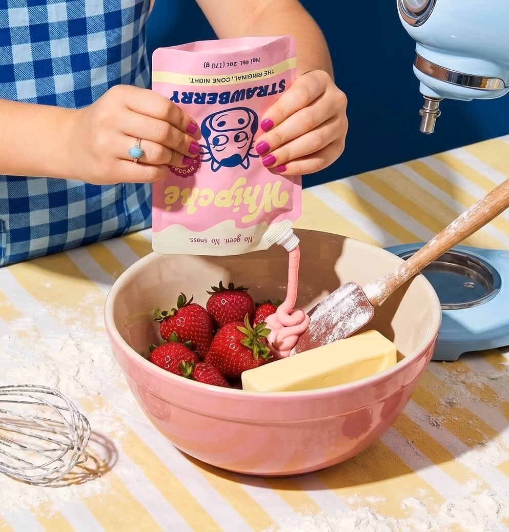 Person in a blue gingham apron squeezes pink strawberry-flavored Whipché cream cheese from a pouch into a mixing bowl filled with fresh strawberries and butter, beside a stand mixer on a baking prep surface.