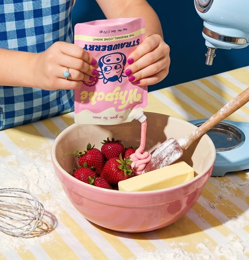 Person in a blue gingham apron squeezes pink strawberry-flavored Whipché cream cheese from a pouch into a mixing bowl filled with fresh strawberries and butter, beside a stand mixer on a baking prep surface.