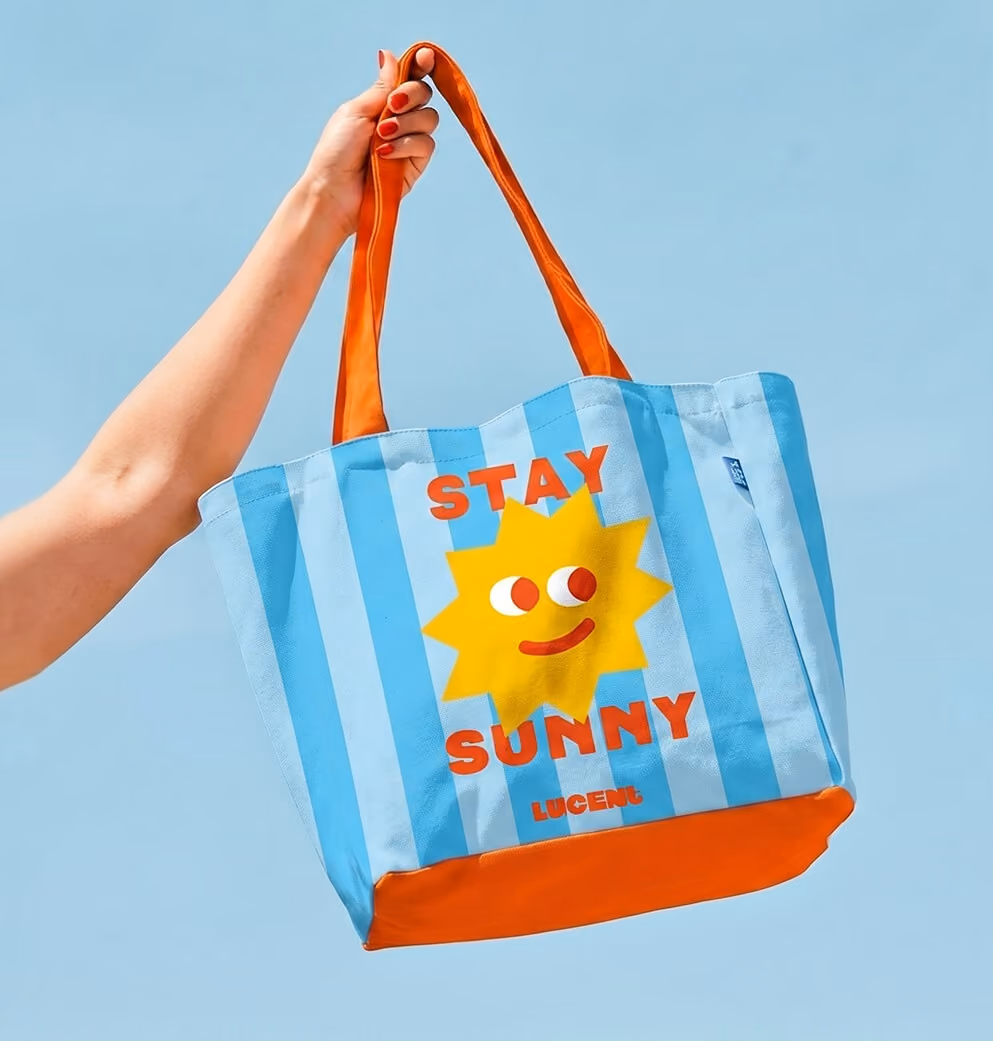 Hand holding a blue-and-white striped Lucent Sun tote bag with orange handles, featuring a smiling sun mascot and the phrase “Stay Sunny” against a clear blue sky.
