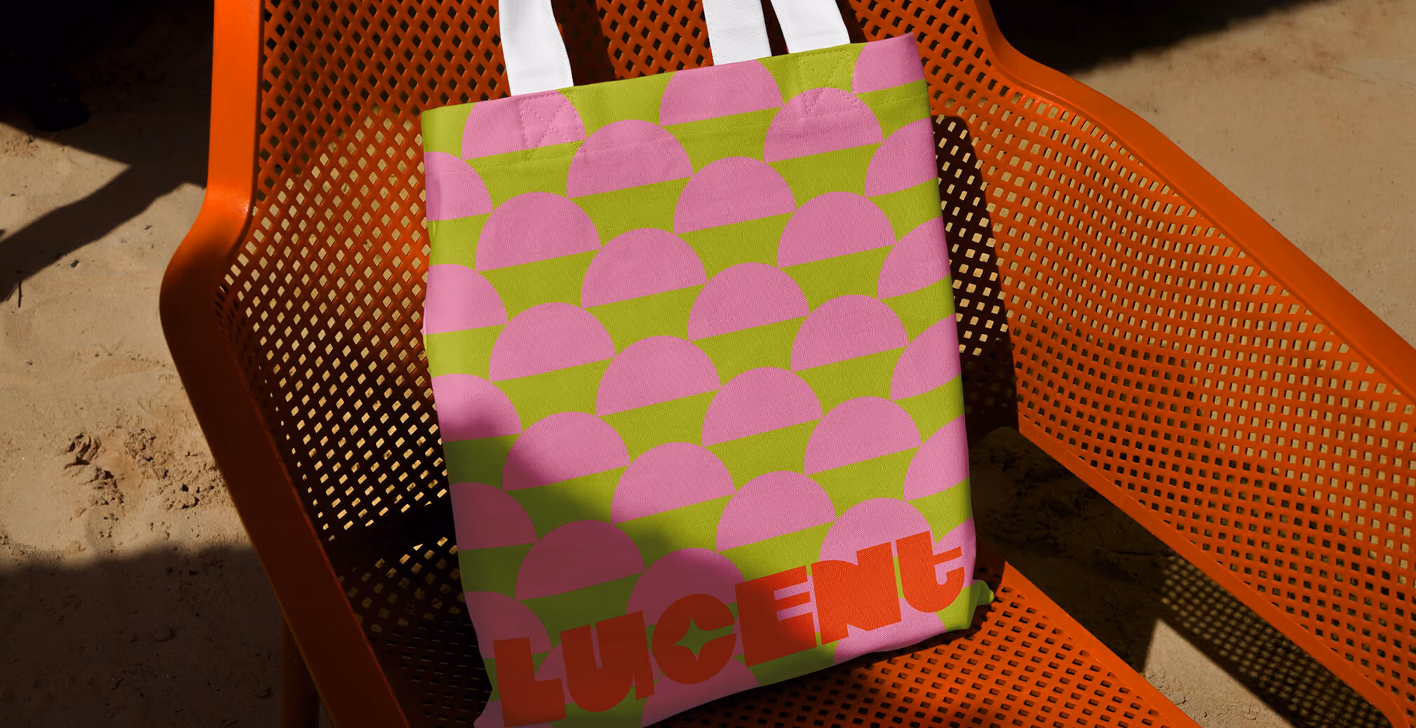 Lucent Sun tote bag featuring a pink and green geometric pattern resting on an orange perforated lounge chair in a sunny outdoor setting.