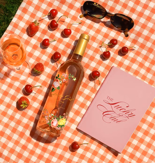 Lucky Girl rosé picnic setup with wine bottle, fruit, sunglasses, and The Lucky Club booklet.