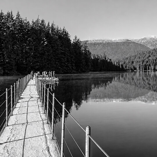 Peaceful lakeside boardwalk in Coquitlam leading across calm water surrounded by evergreen forests and mountain scenery.