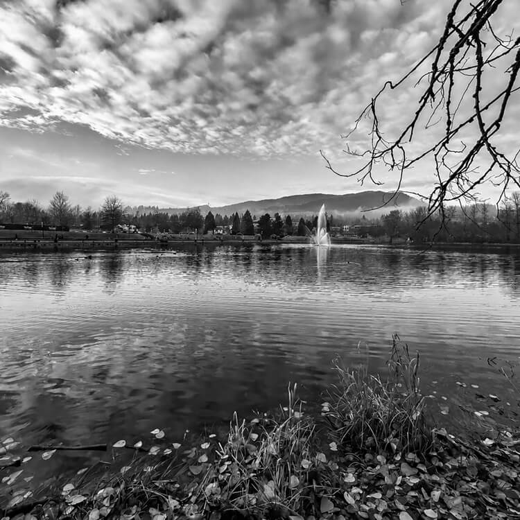 Lafarge Lake in Coquitlam with a central fountain, reflective waters, and mountain views beneath textured clouds.