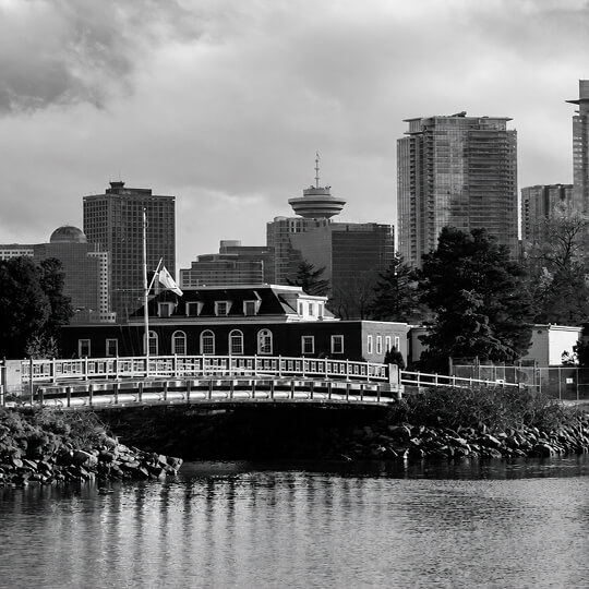 New Westminster waterfront with a pedestrian bridge, historic riverside architecture, and the Metro Vancouver skyline in the background.