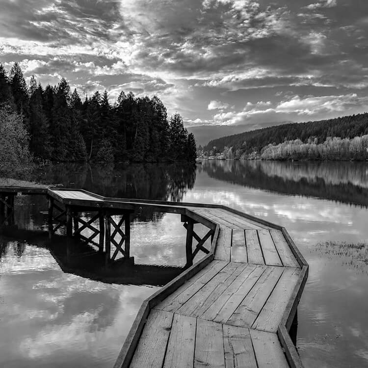 Wooden boardwalk extending over a calm lake surrounded by dense evergreen forest and reflected clouds.