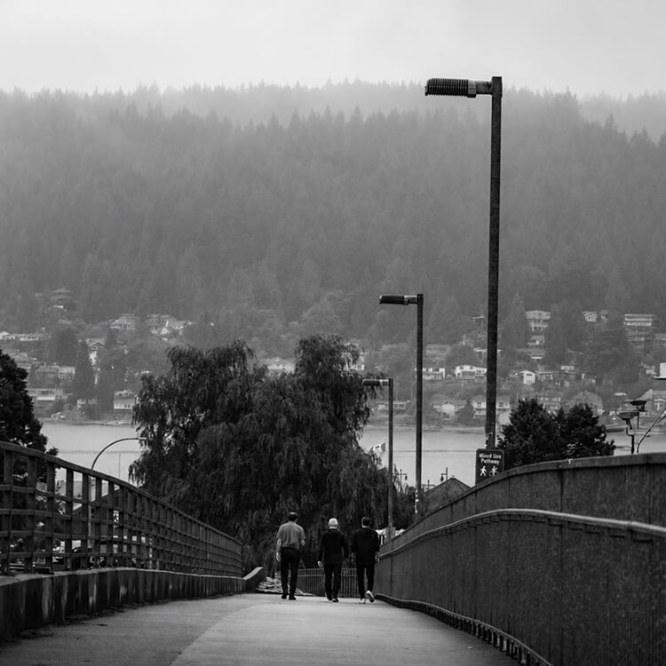 Pedestrians walking along a pathway overlooking the Port Moody waterfront and forested hillside neighbourhoods.