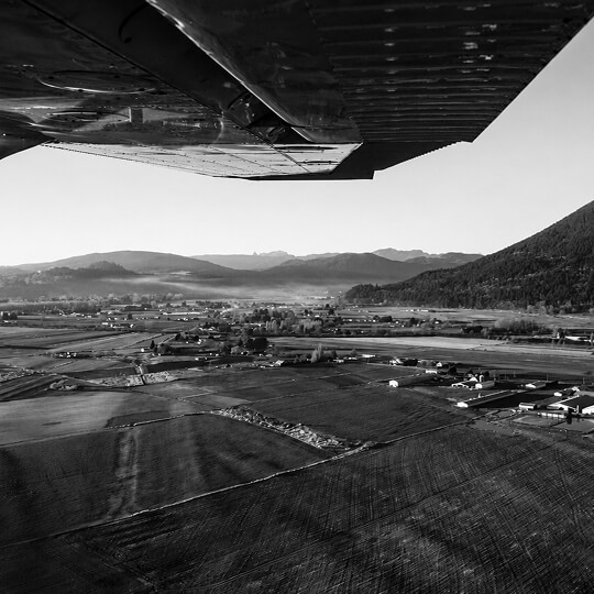 Black-and-white aerial view from beneath an airplane wing, overlooking rural farmland, roads, and distant mountains around Langley.