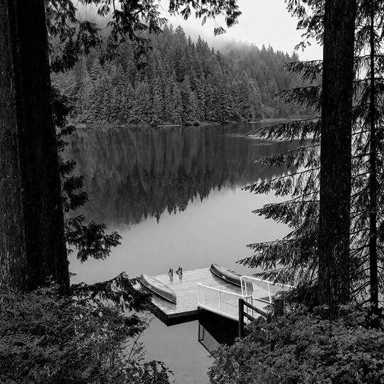Black-and-white view of a peaceful forest lake with a small dock and canoes surrounded by dense evergreen trees.