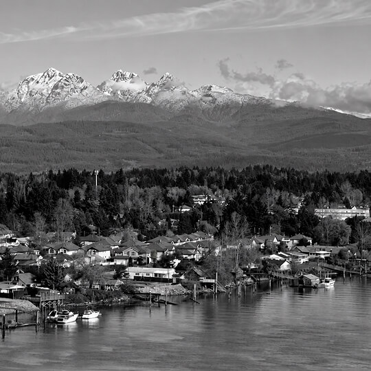 Black-and-white waterfront neighborhood in Maple Ridge with homes, boats, forested hills, and snow-covered mountains in the distance.