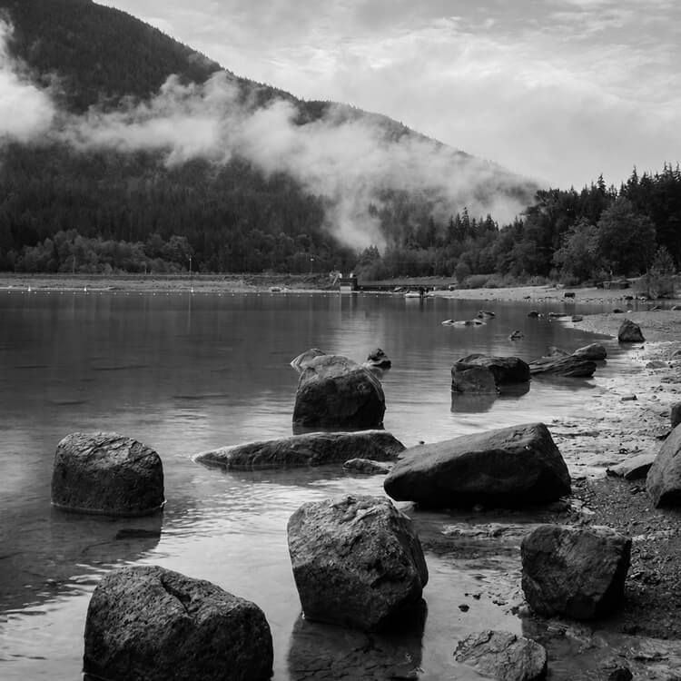 Black-and-white rocky lakeshore with calm water, forested mountains, and low clouds drifting across the landscape.