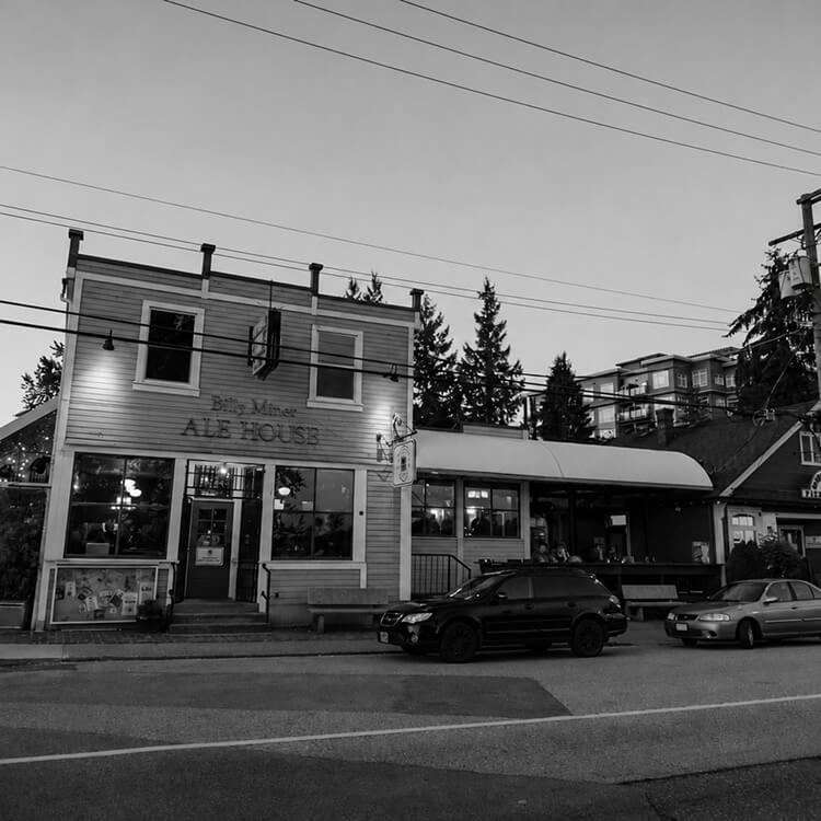 Black-and-white street view of a local Maple Ridge ale house with parked cars, storefronts, and surrounding buildings.