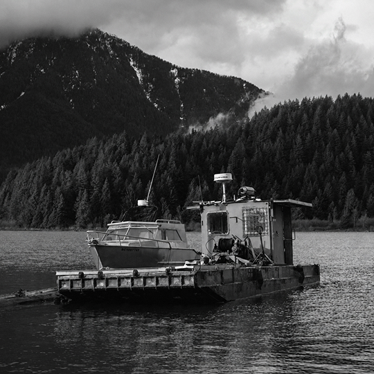 Workboats rest on calm water beneath forested mountains and low clouds near Pitt Meadows, British Columbia.