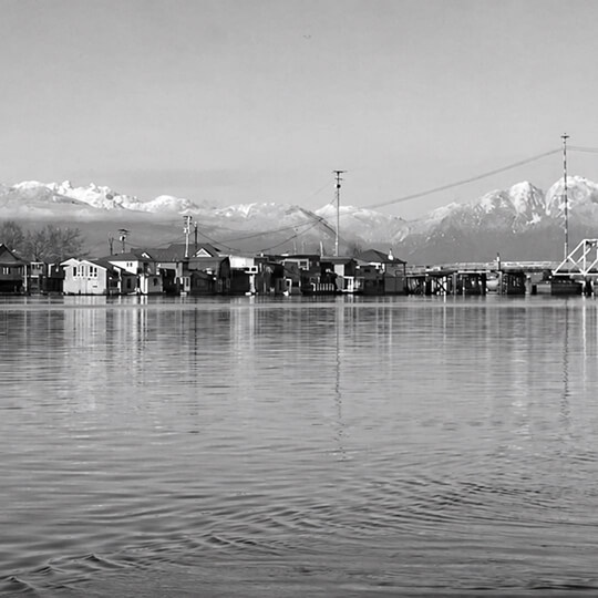 Waterfront homes sit along calm water with snow-capped mountains rising in the distance near Delta, British Columbia.