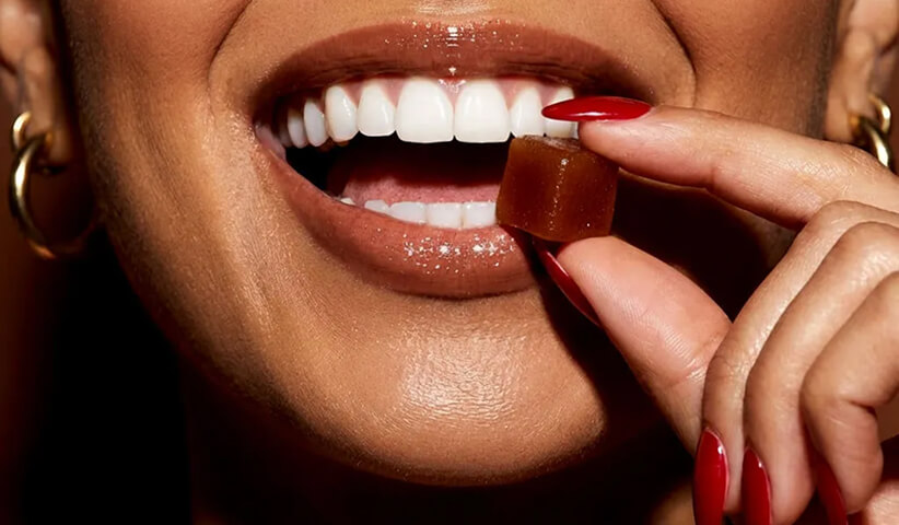 Close-up of a woman holding a brown gummy near her mouth with red nails and gold hoop earrings.