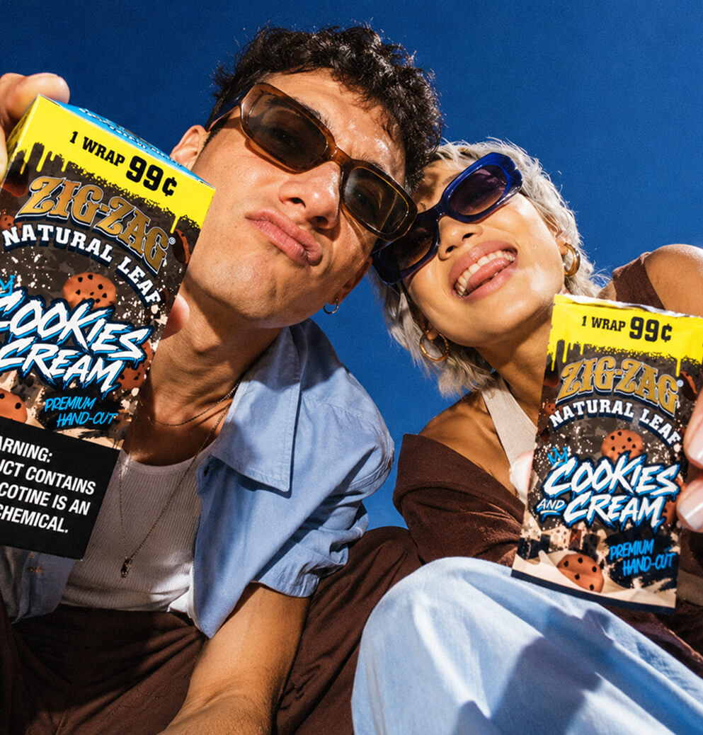 Two people wearing sunglasses pose with Zig-Zag Natural Leaf Cookies and Cream wrap packages against a vivid blue sky.