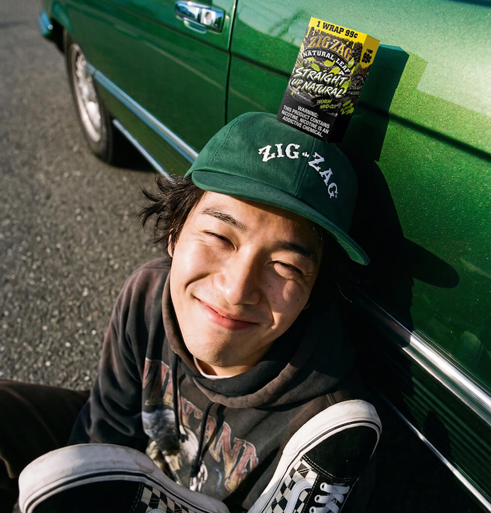 Smiling person wearing a green Zig-Zag cap sits beside a green car with a Straight Up Natural wrap package displayed above them.