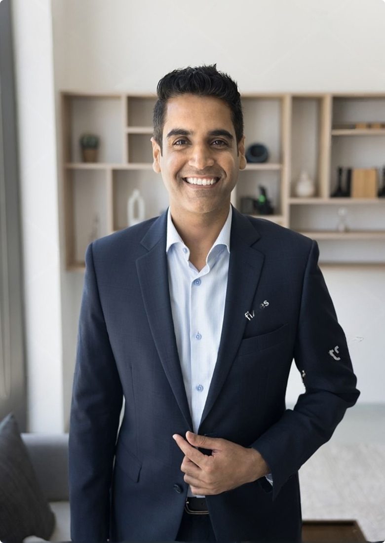 Smiling man in a dark blue suit jacket and light blue shirt stands indoors with shelves and decor in the background.