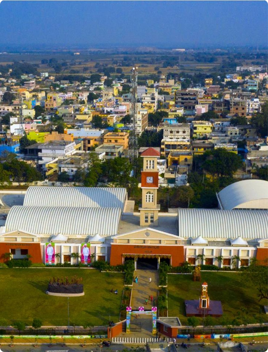 Aerial view of a large building with a clock tower, surrounded by green lawns and cityscape in the background.