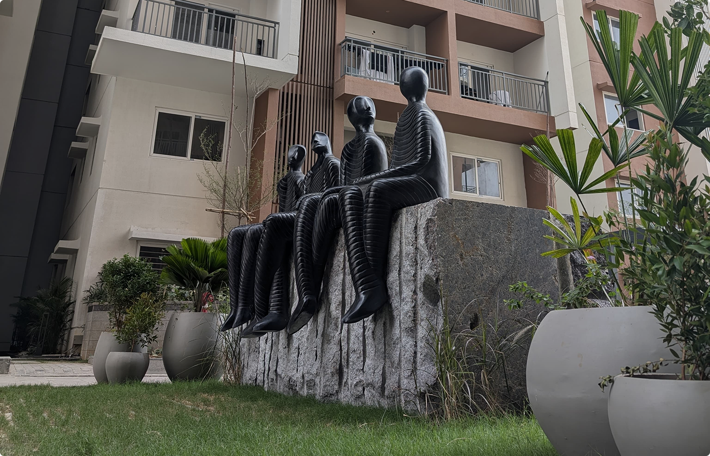 Four stylized black human sculptures with ribbed texture sitting on a large stone block in a garden area in front of a modern building.