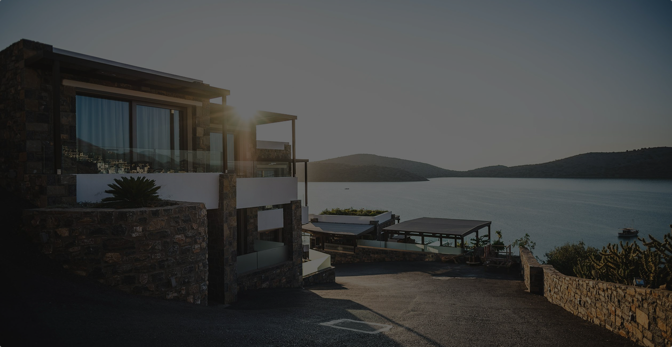 Sunset view of a modern stone house overlooking a calm sea with hills in the background.