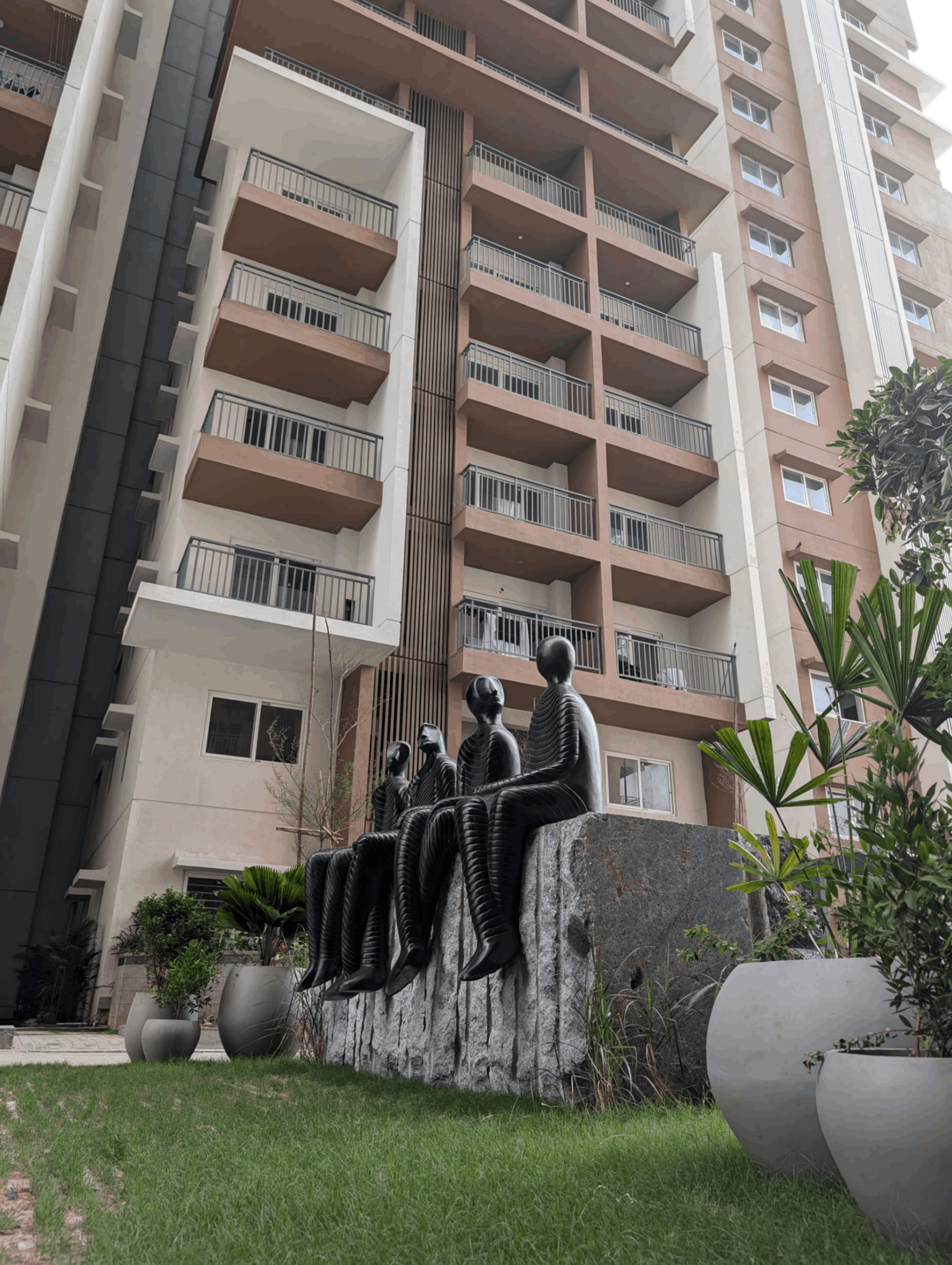 Four black abstract human statues sitting on a large stone block in front of a multi-story residential building with balconies.