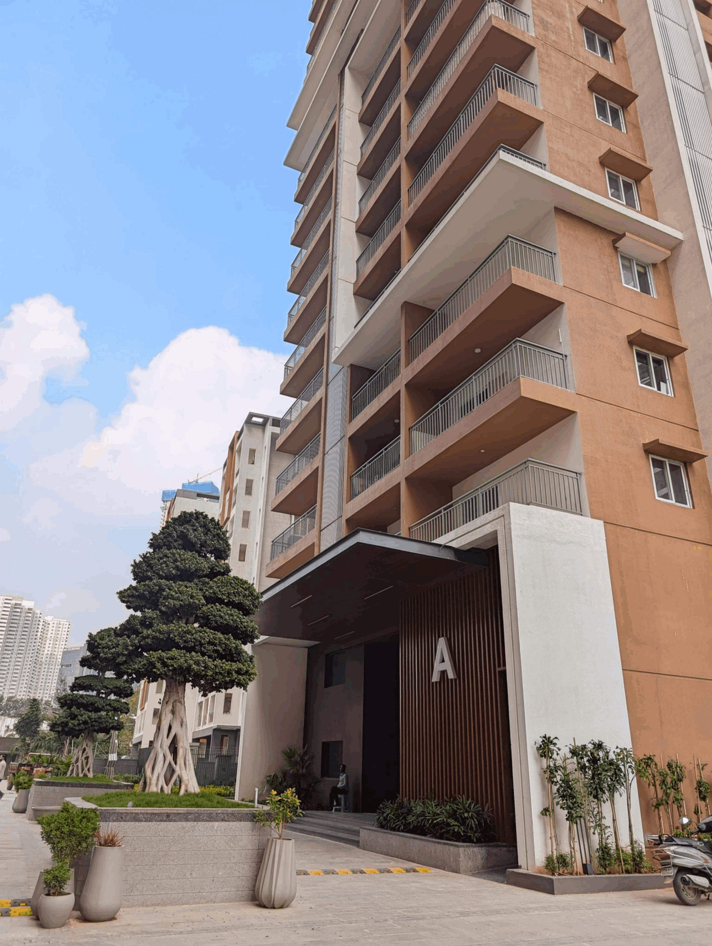 Modern multi-story apartment building with balconies, landscaped entrance marked with letter A, and well-maintained trees and plants.