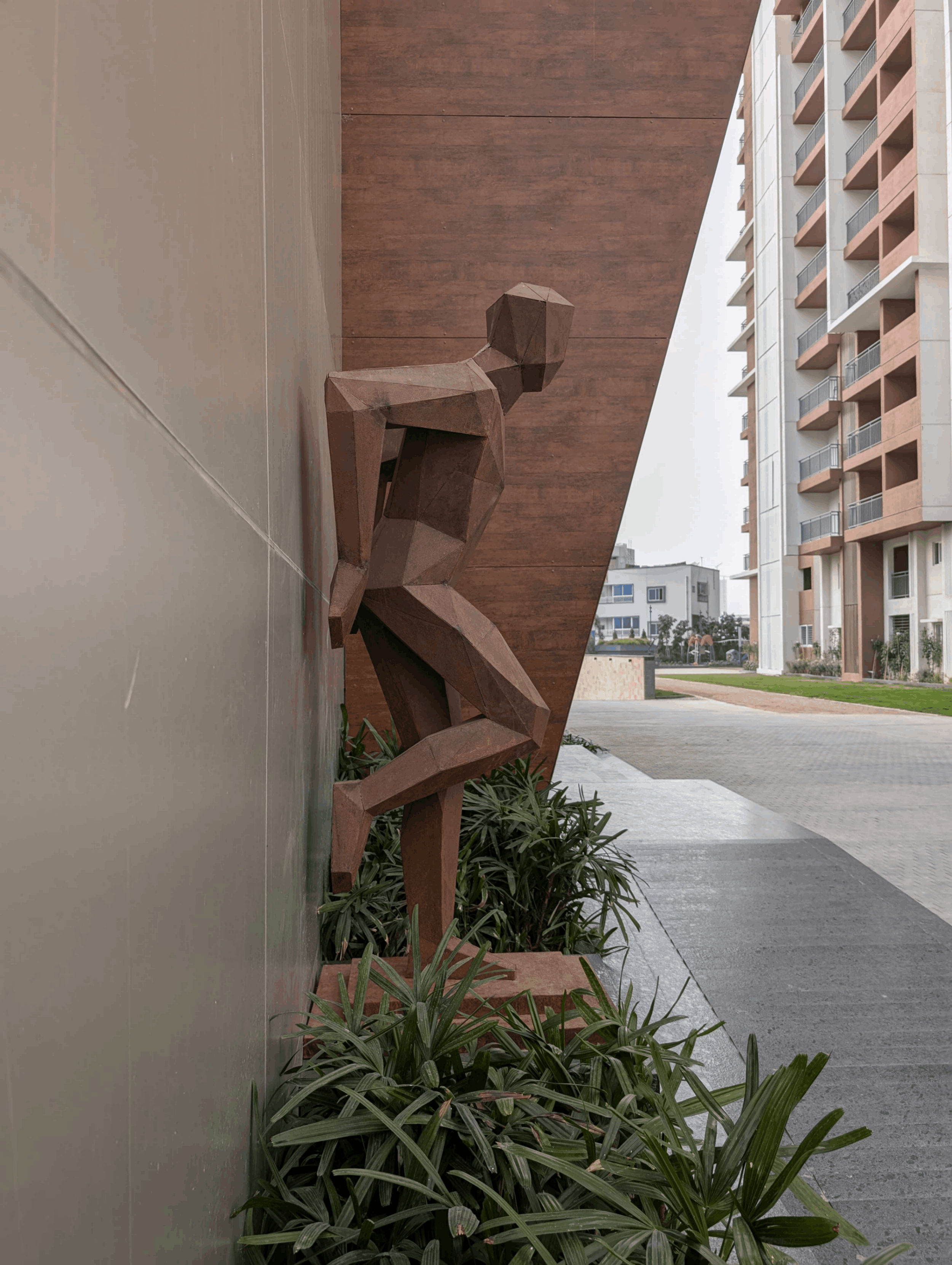 Geometric metal sculpture of a human figure leaning against a wall next to green plants, with modern buildings in the background.