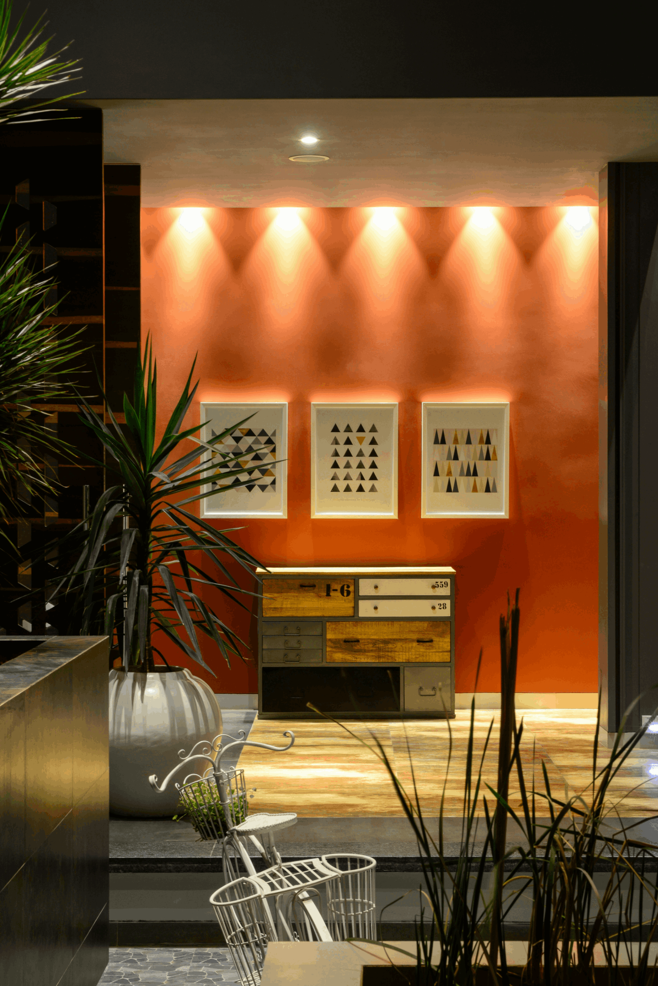 Indoor space with warm orange wall, three framed geometric art pieces, a multi-drawer wooden chest, and green plants in white pots.