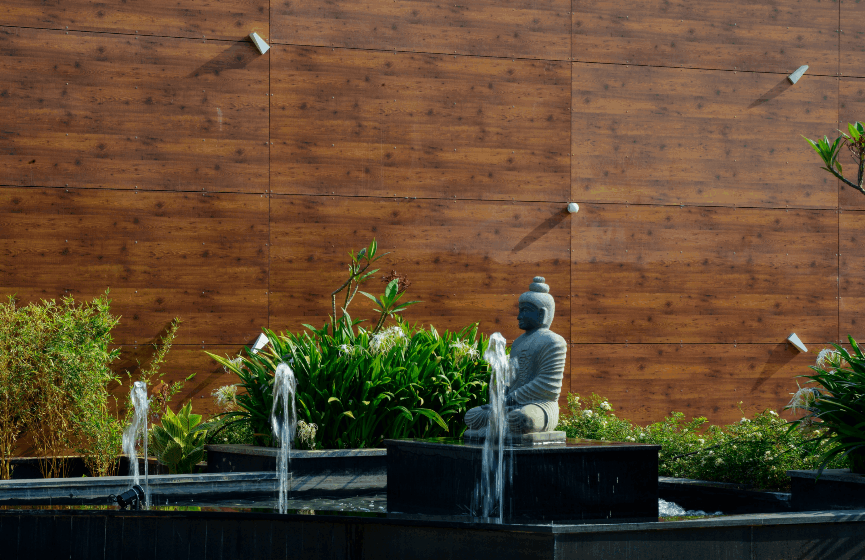 Stone Buddha statue sitting on a pedestal in a water fountain surrounded by green plants and a wooden wall background.