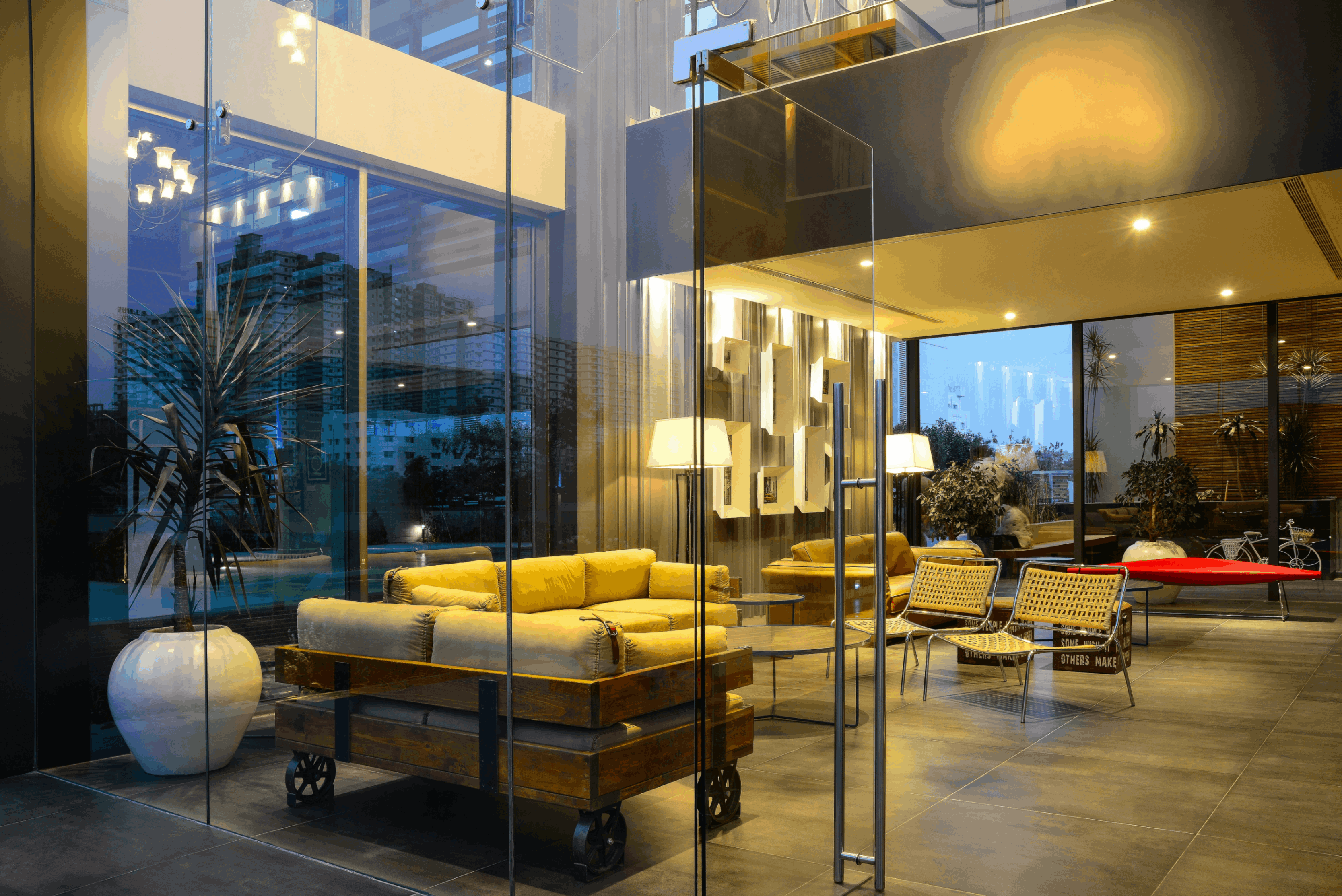 Modern indoor seating area with wooden sofas, woven chairs, large potted plants, and glass walls showing city buildings at dusk.