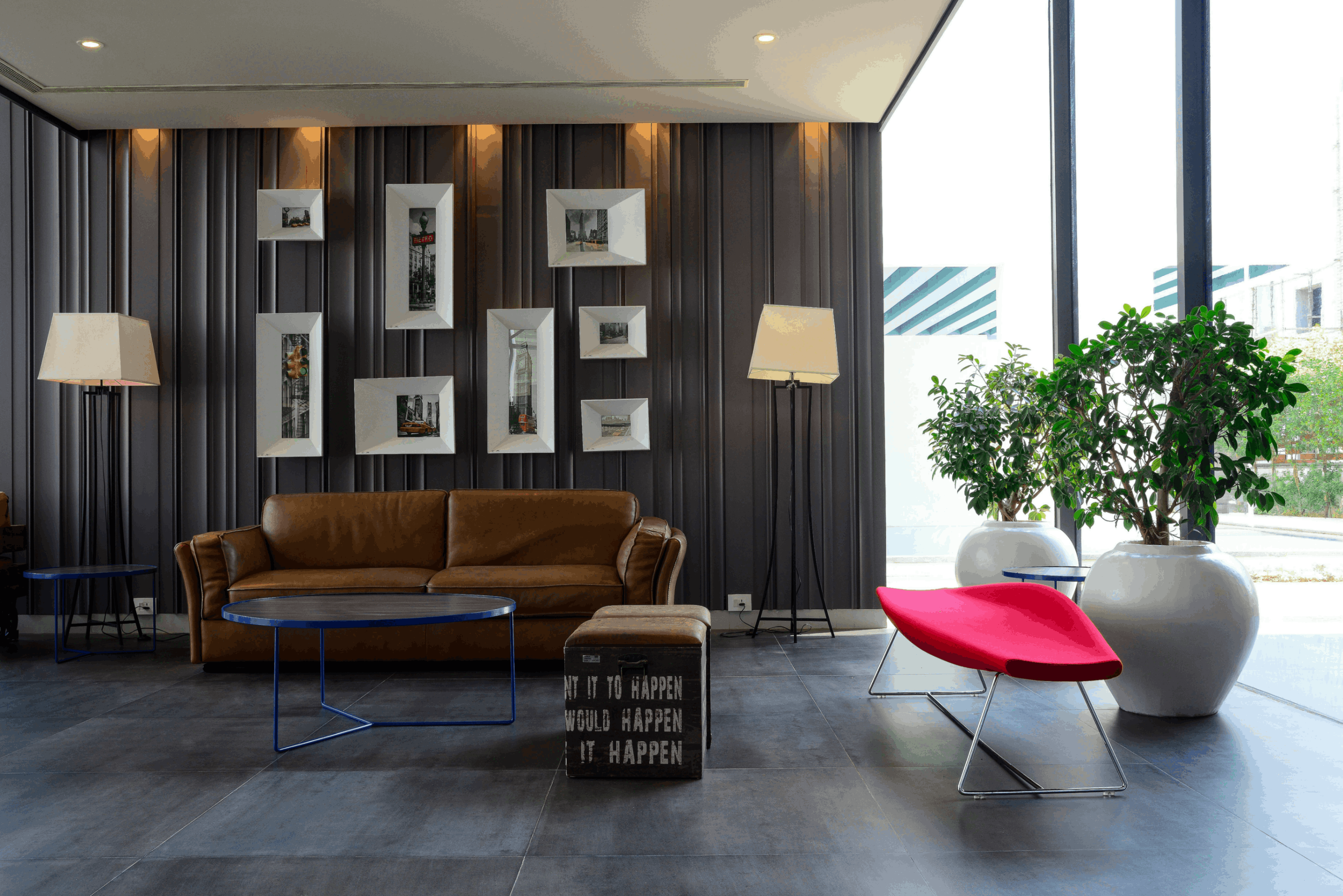 Modern living room with a brown leather sofa, blue metal coffee table, red accent chair, large white pots with green plants, and framed black-and-white city photos on a dark wood-paneled wall.