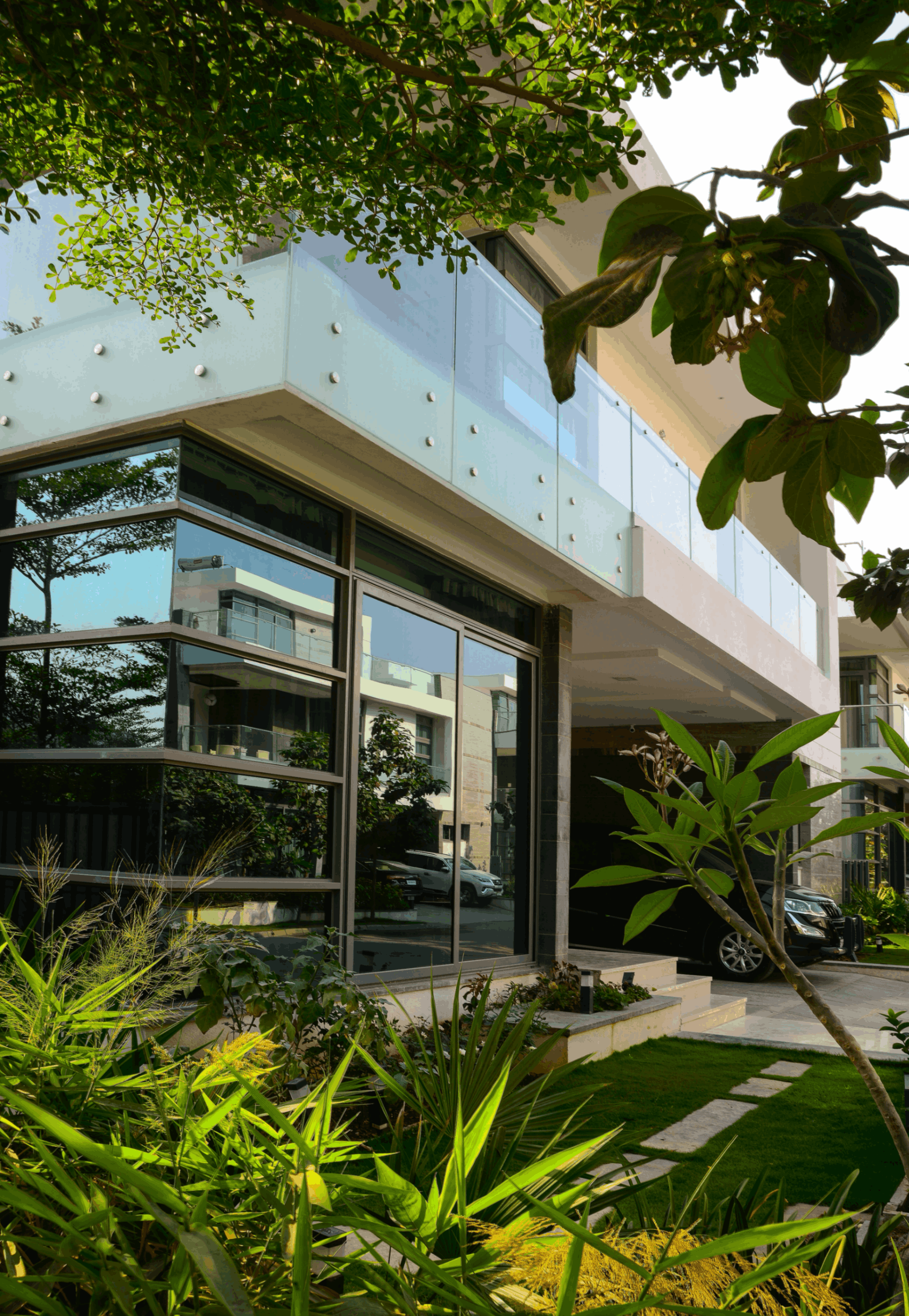 Modern house with glass balcony, large reflective windows, green plants, and parked cars nearby.