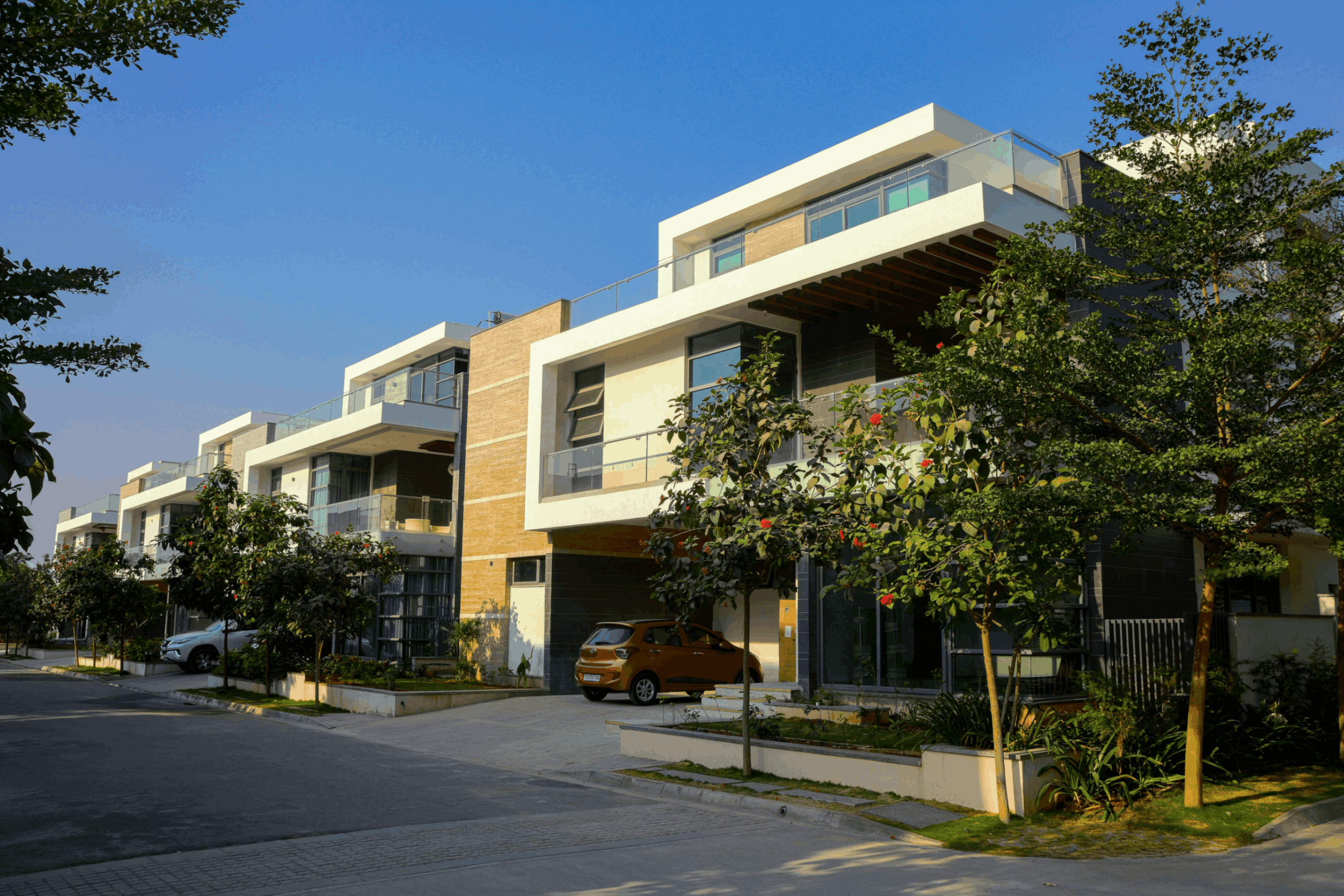 Modern multi-story residential buildings with large windows and balconies lining a street with trees and parked cars under a clear blue sky.
