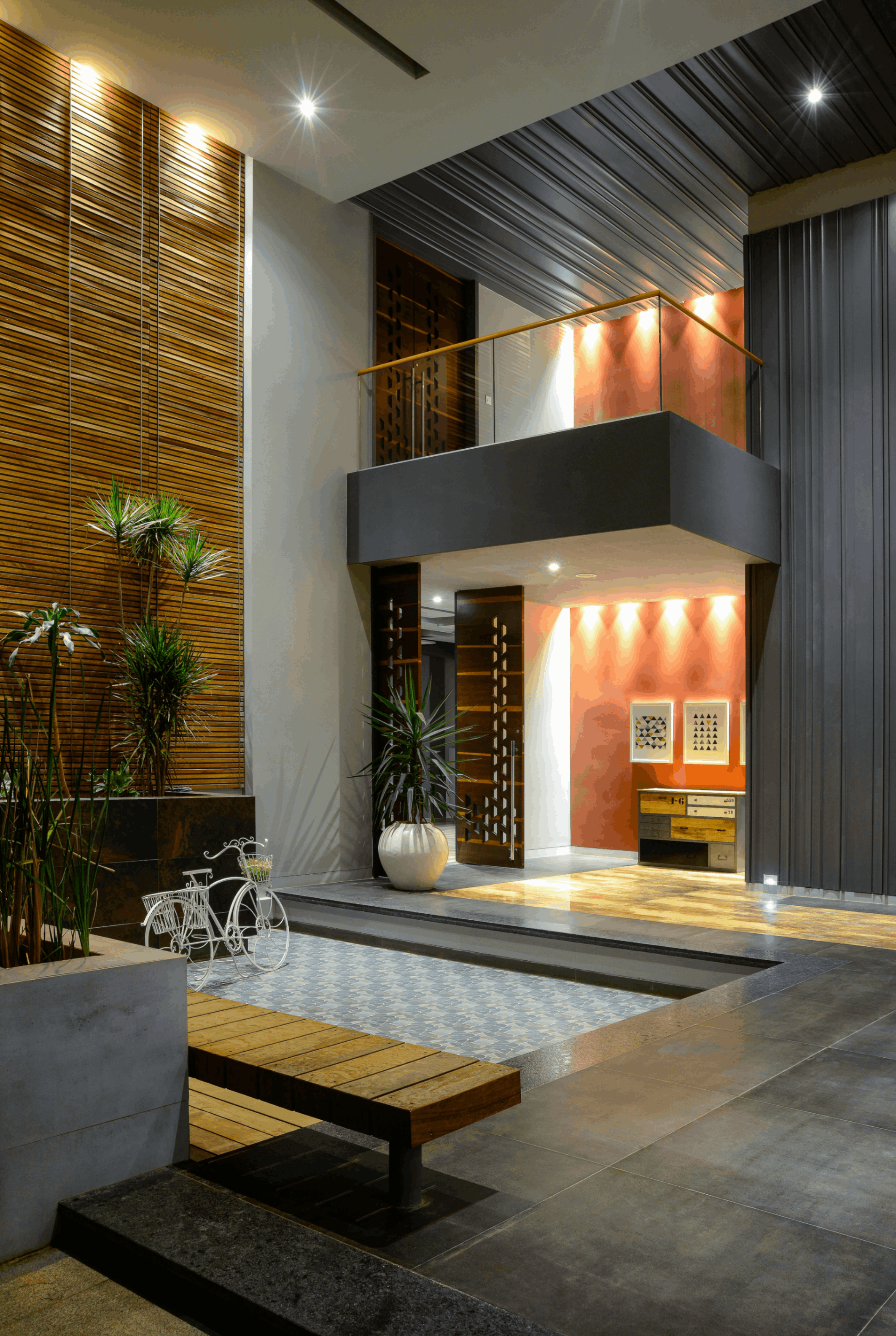 Modern house entrance with wood paneling, large potted plants, a decorative white bicycle, wood bench, tiled shallow pool, and warm indoor lighting.