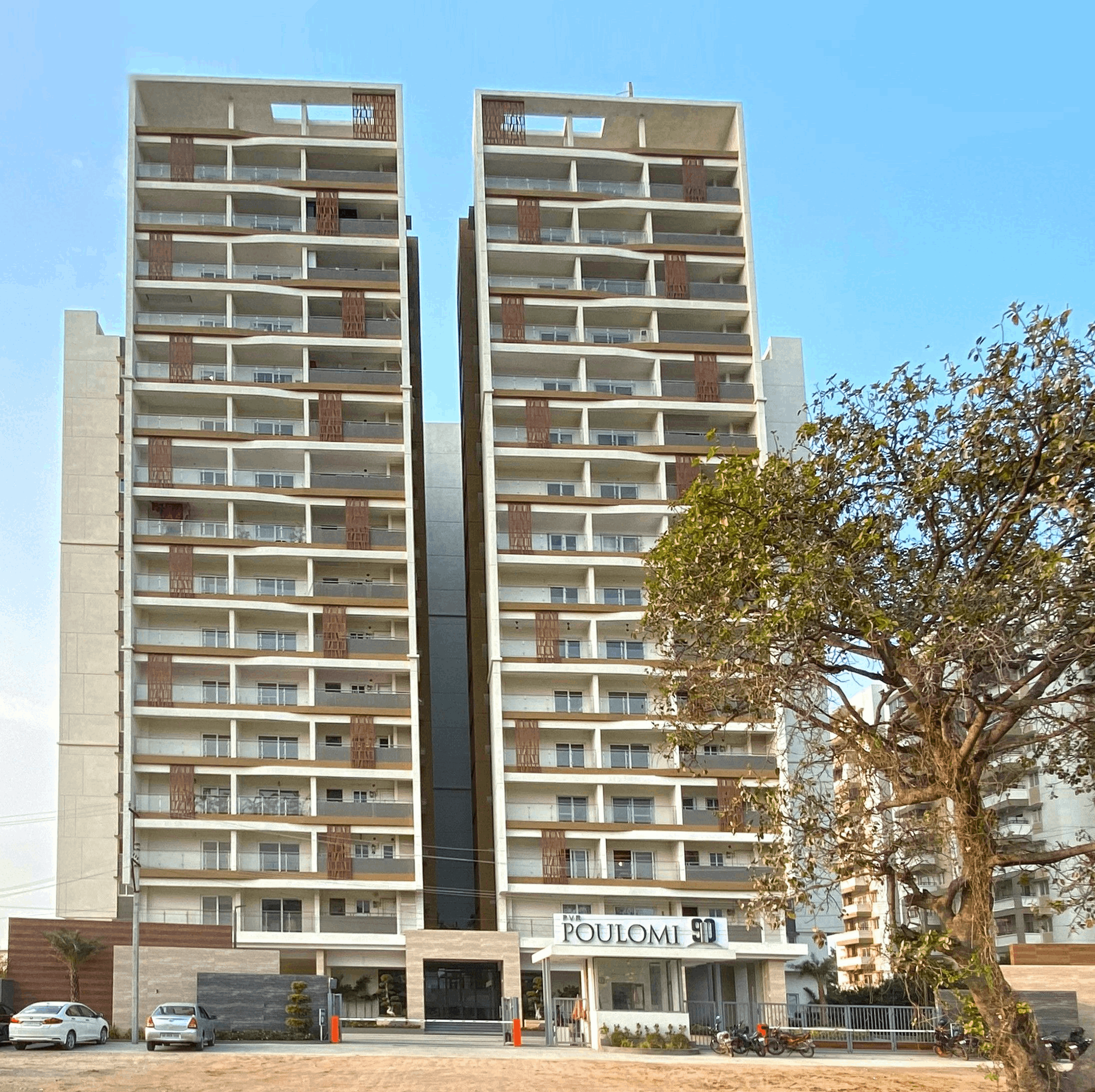 Two modern beige residential buildings labeled 'Poulomi 90' with balconies and a gated entrance, a tree on the right, and parked cars and motorcycles in front.
